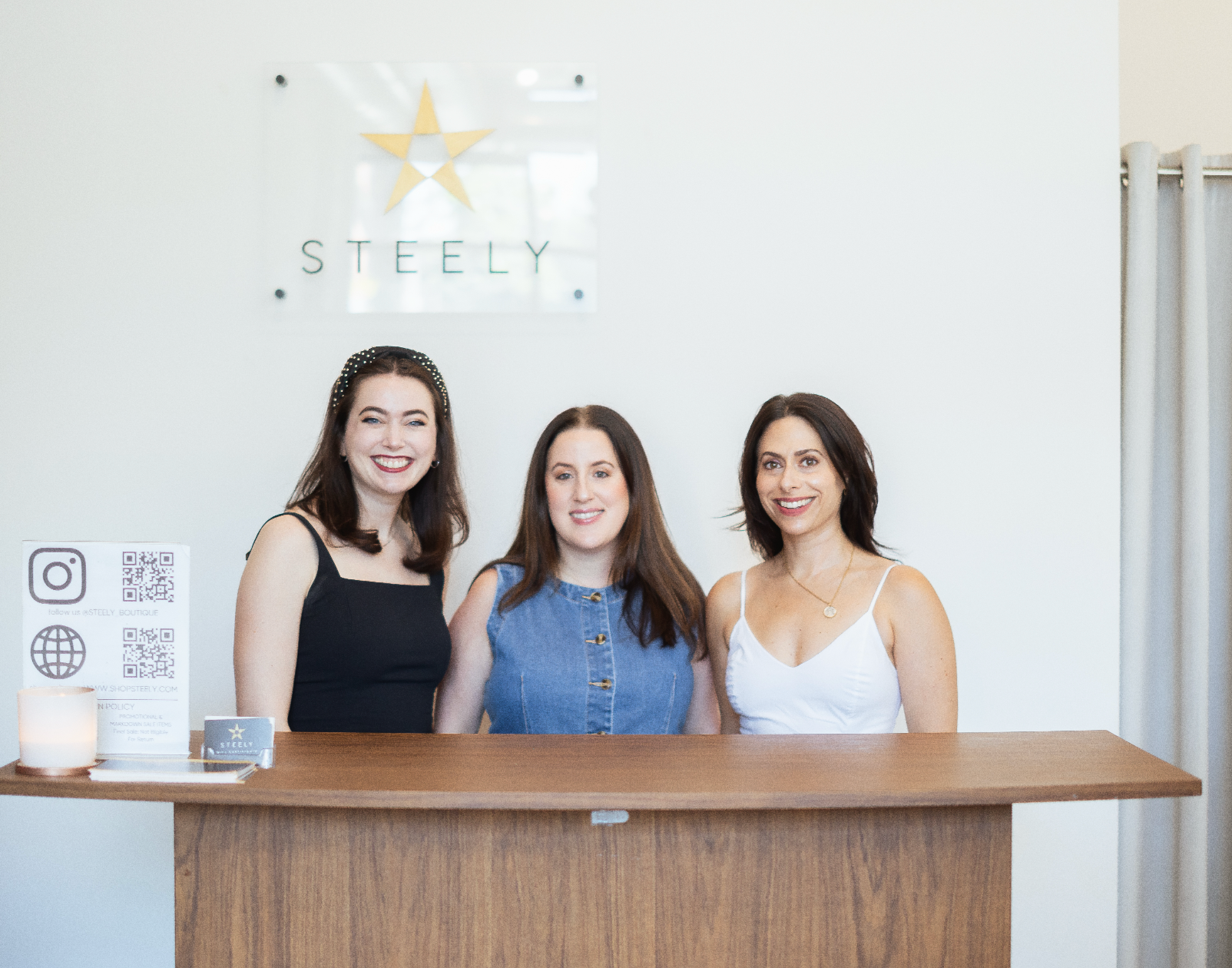 Three women standing behind a reception desk with 'Steely' branding in the background.
